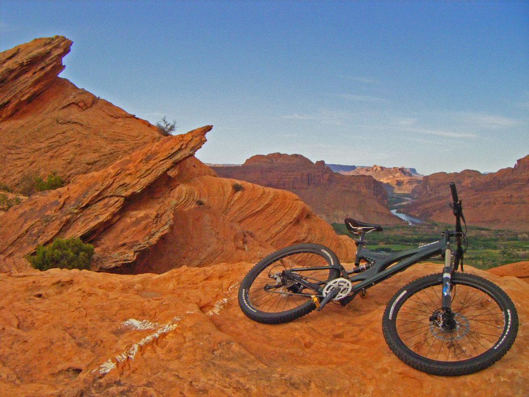 A mountain bike resting on a rocky red landscape, overlooking a winding river and canyon in the background, under a clear blue sky. Slickrock mountain bike trail.