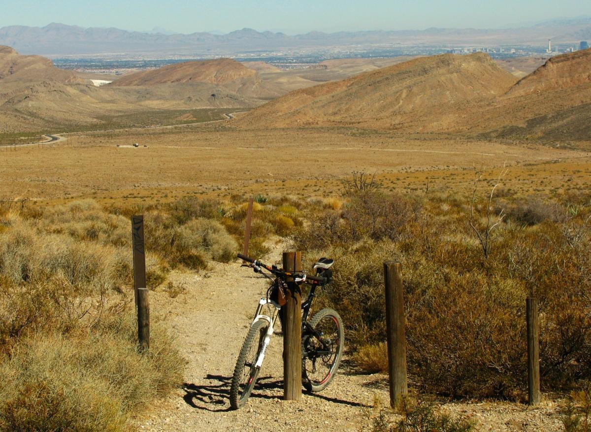 A mountain bike parked near a trail marker, overlooking a vast desert landscape with hills and distant mountains. The scene shows a clear blue sky and dry terrain with sparse vegetation. Deadhorse Loop mountain bike trail.
