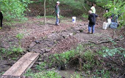 A small wooden bridge crosses over a shallow stream in a forested area. Several people are engaged in outdoor work, standing on the rocky path and near buckets in the background. Lush green foliage surrounds the scene, indicating a natural setting. North Park mountain bike trail.