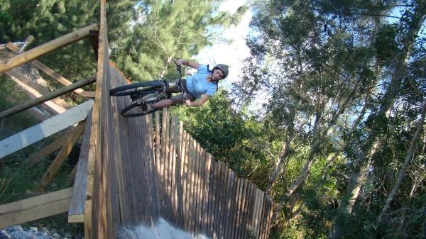 A person riding a mountain bike on a wooden ramp in a forested area, leaning to the side while navigating the curve of the ramp. Sunlight filters through the trees in the background. The cyclist is wearing a helmet and casual clothing. Markham Park mountain bike trail.