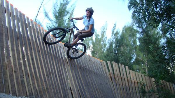 A person riding a mountain bike performs a trick on a wooden ramp, leaning into the turn as they navigate a steep incline. The scene is set outdoors with trees in the background and a clear blue sky. The rider is wearing a helmet and casual clothing. Markham Park mountain bike trail.
