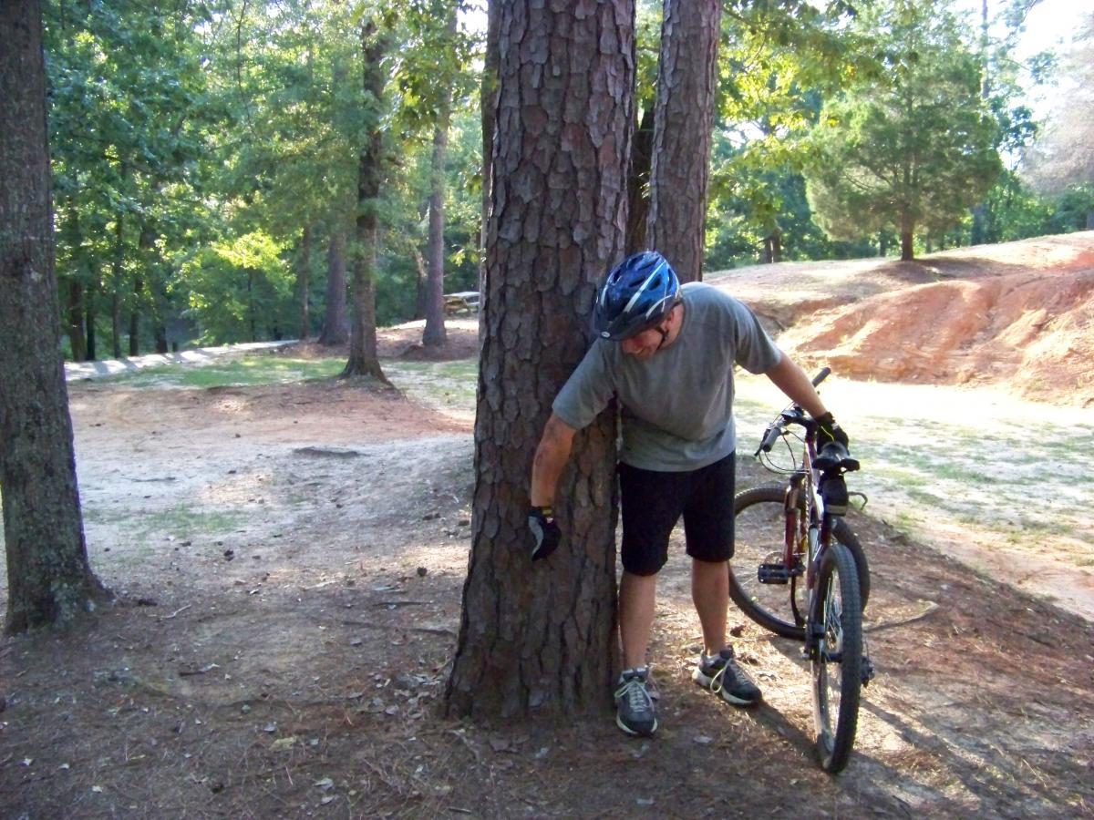 A person in a helmet stands beside a tree in a wooded area, holding onto the tree with one hand and leaning slightly forward. A mountain bike is positioned next to them on the ground. The setting features a path and grassy terrain surrounded by tall trees, suggesting an outdoor biking trail. Flat Rock Park mountain bike trail.