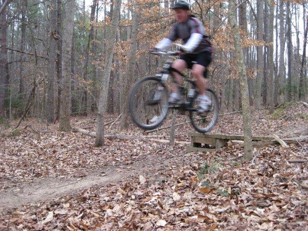 A mountain biker in action, jumping off a small wooden ramp on a trail surrounded by trees and autumn leaves. The cyclist is wearing a helmet and athletic gear, with the bike lifted off the ground as he prepares to land. Freedom Park (aka:district Park) mountain bike trail.