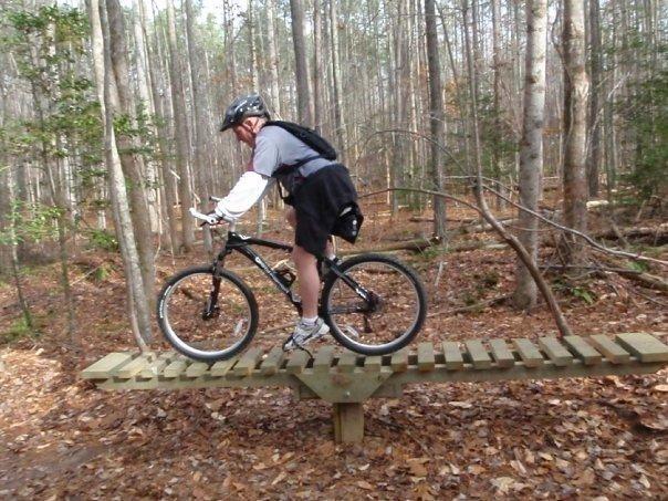 A cyclist navigating a narrow wooden bridge while mountain biking in a forested area, surrounded by trees and fallen leaves. The rider is wearing a helmet and biking gear, showing focus and balance on the trail. Freedom Park (aka:district Park) mountain bike trail.