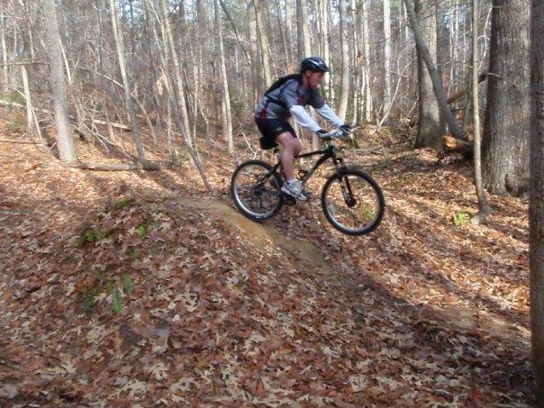 A mountain biker in a helmet and athletic gear is in mid-air, jumping off a small dirt mound on a forest trail covered with autumn leaves. Surrounding trees have sparse foliage, indicating a late fall season. Freedom Park (aka:district Park) mountain bike trail.