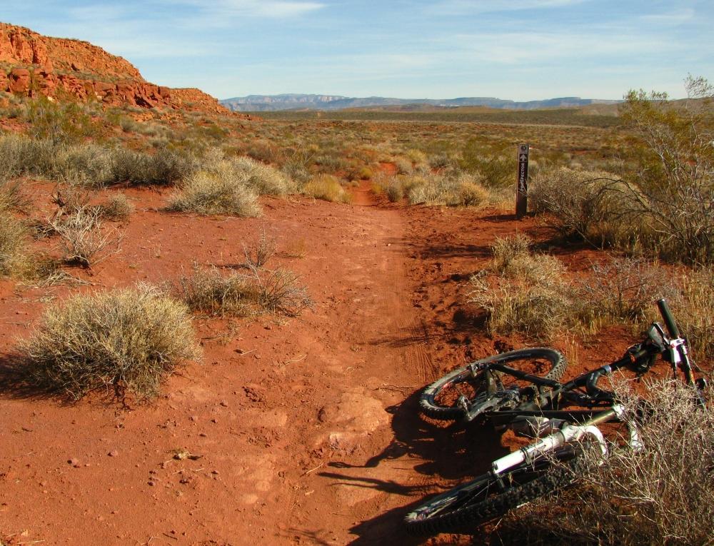 A mountain bike lying on a dirt path surrounded by sparse vegetation and red rocky terrain, with distant mountains in the background and a signpost indicating a trail. The scene is set in a sunny, clear day. Prospector - Church Rocks Loop mountain bike trail.