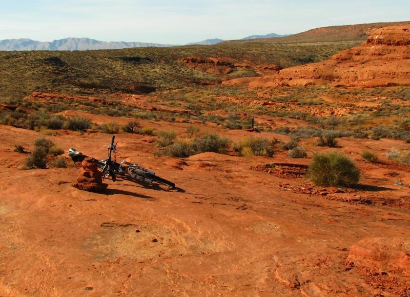 A mountain bike is leaning against a stack of rocks on a rocky, red terrain. The landscape features sparse vegetation and rolling hills, under a clear blue sky with distant mountains in the background. Prospector - Church Rocks Loop mountain bike trail.