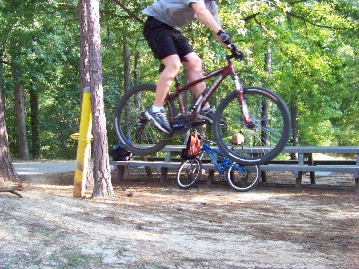 A mountain biker in mid-air performing a jump over a log, with trees and a picnic area visible in the background. A blue bike can be seen resting on the ground nearby. Flat Rock Park mountain bike trail.