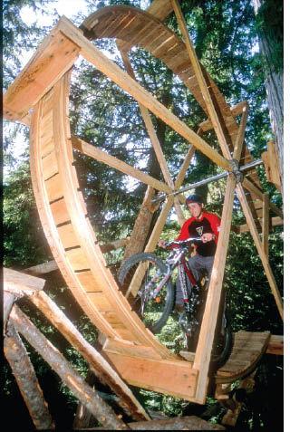 A mountain biker riding on a large wooden wheel structure built into a forested environment, surrounded by tall trees. The biker is wearing a helmet and a red jersey, focusing on navigating the unique wooden feature.