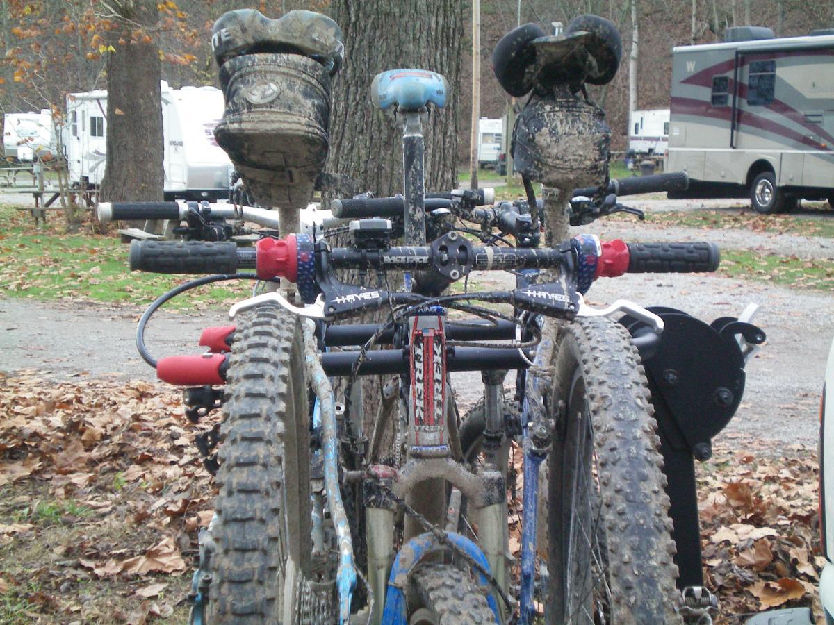 Close-up view of several mountain bikes with mud-covered tires and handlebars, lined up in a wooded area, with recreational vehicles visible in the background among fallen leaves. Carvin's Cove Trail system mountain bike trail.