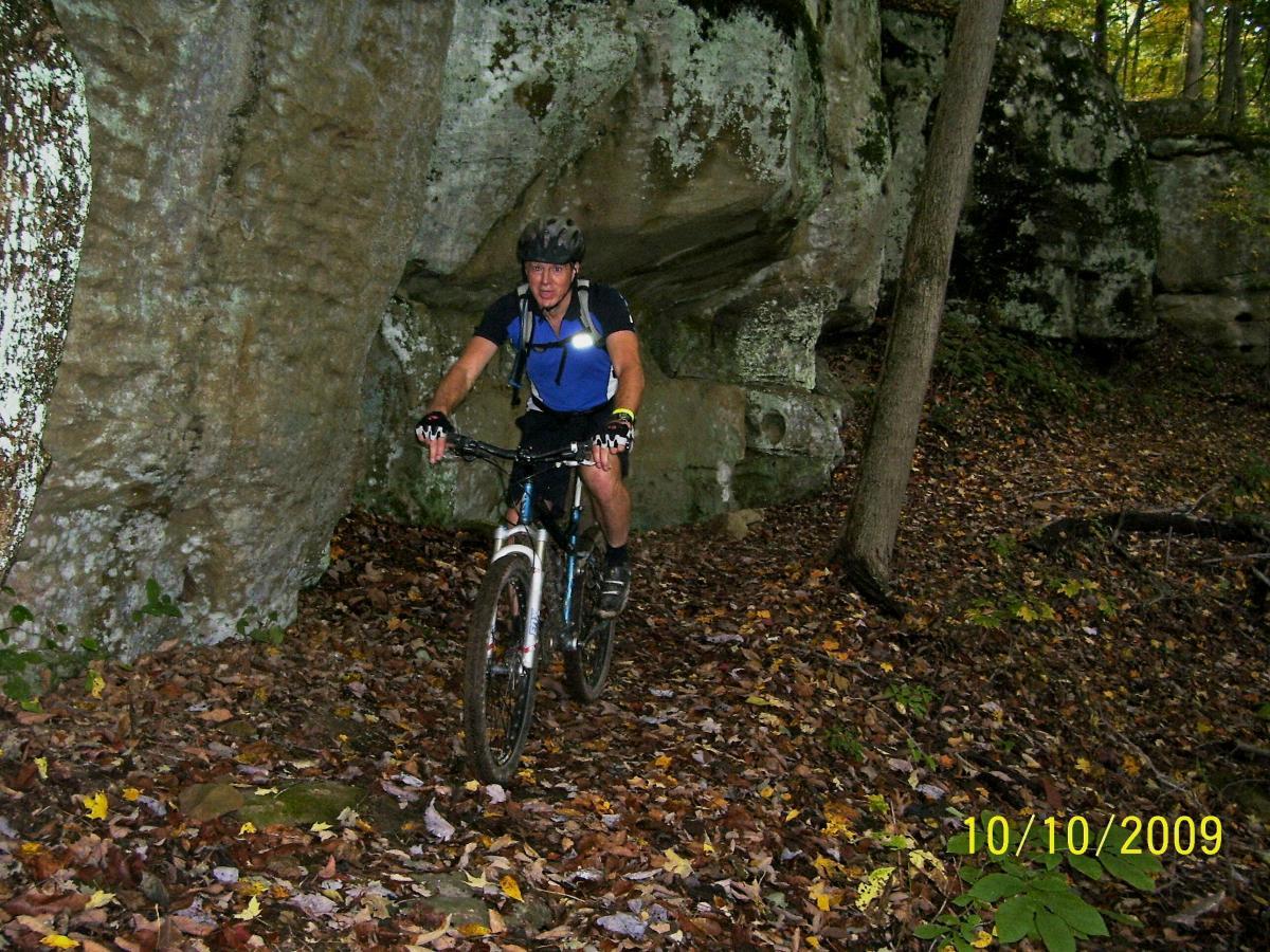A mountain biker rides on a narrow trail surrounded by autumn foliage and rocky terrain. The cyclist is wearing a helmet and athletic clothing, navigating through a wooded area with large rock formations in the background. Fallen leaves cover the ground, indicating a seasonal backdrop. Ohio View Trail mountain bike trail.