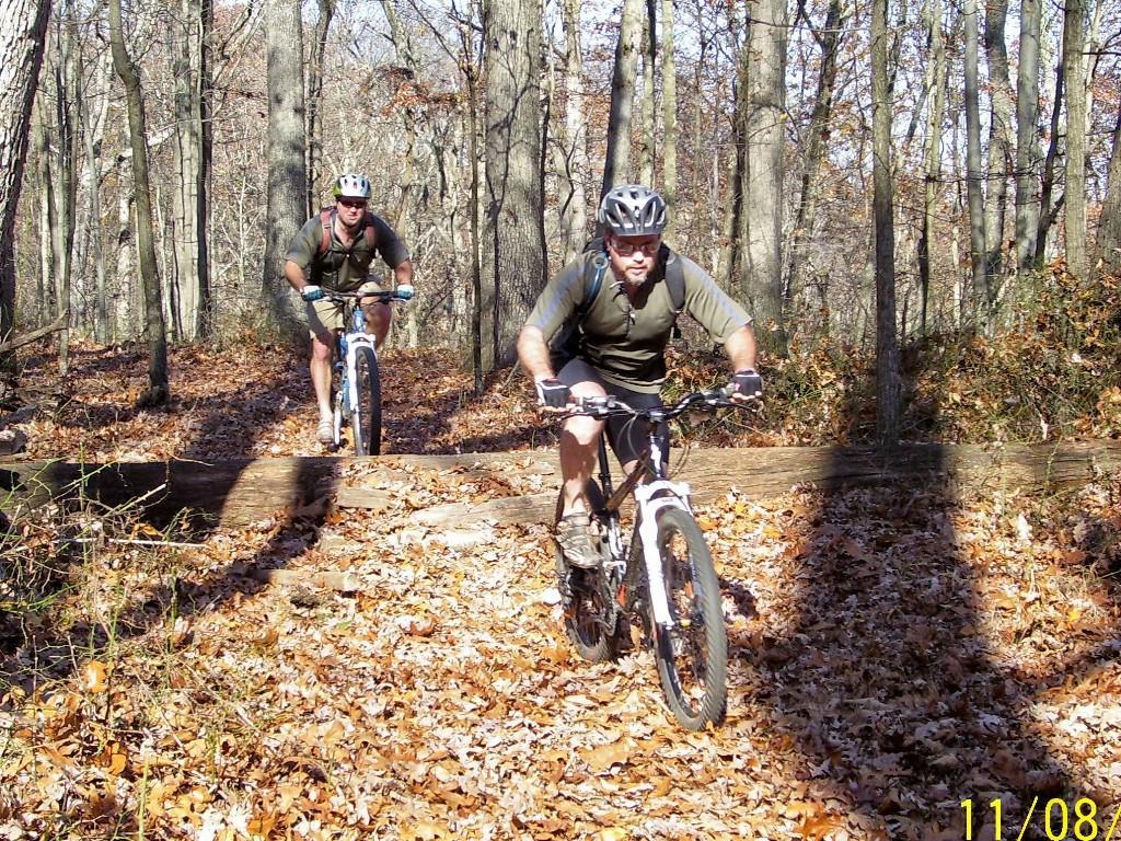 Two mountain bikers navigate a wooded trail in autumn, surrounded by trees and fallen leaves. The first biker, in the foreground, is riding over a log on the path while the second biker trails behind slightly, both wearing helmets and athletic gear. The scene captures the essence of outdoor adventure and cycling. Lake Hope State Park mountain bike trail.