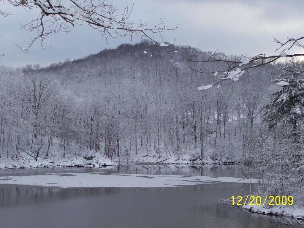 A serene winter landscape featuring a calm river reflecting the surrounding snow-covered trees and a distant hill. The scene is tranquil, with a cloudy sky, and the ground is blanketed in fresh snow, creating a peaceful atmosphere. Mountwood mountain bike trail.