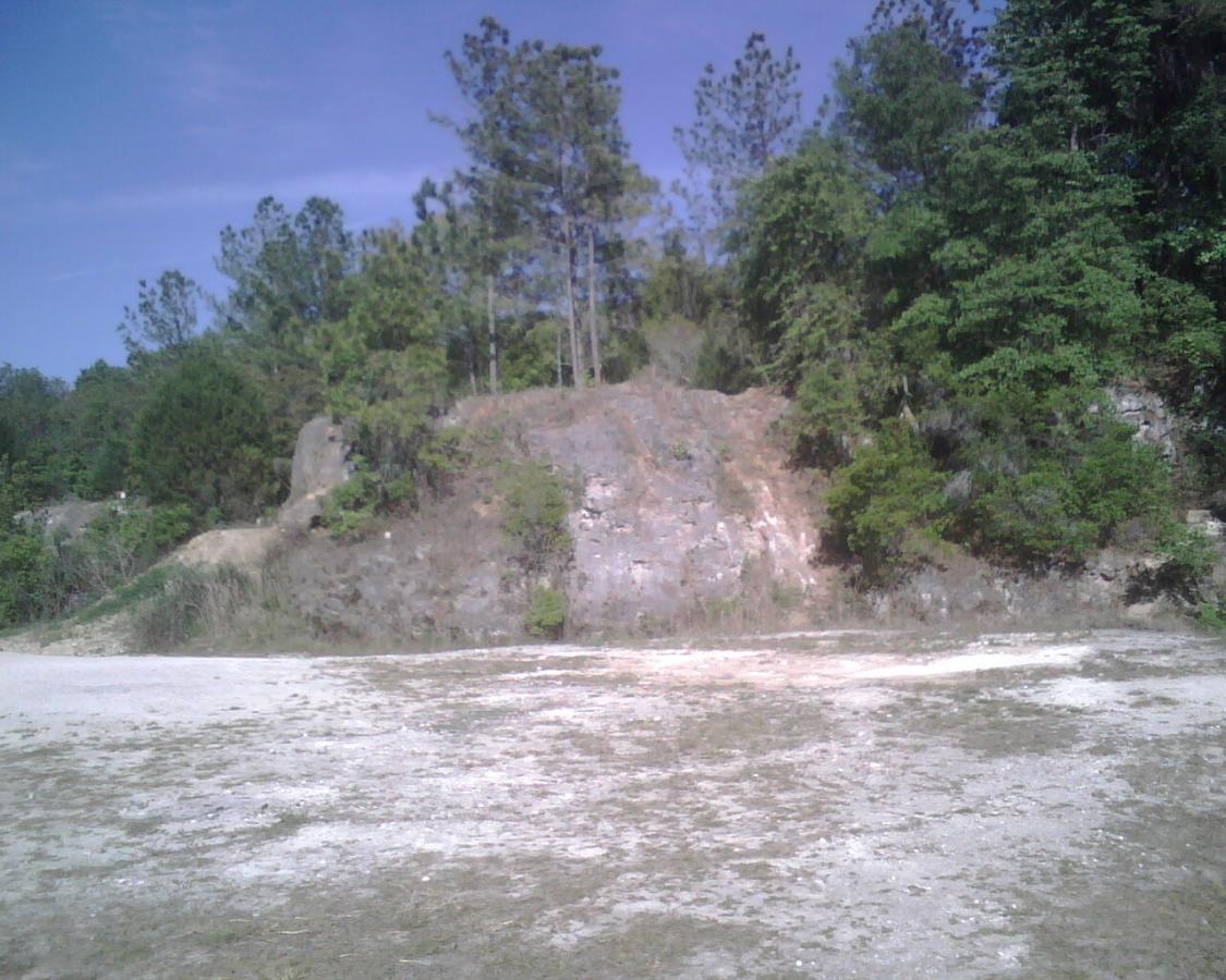 A scenic view of a sloped, rocky area surrounded by green trees under a blue sky. The terrain is mostly bare with patches of dried grass and a few shrubs. Santos mountain bike trail.