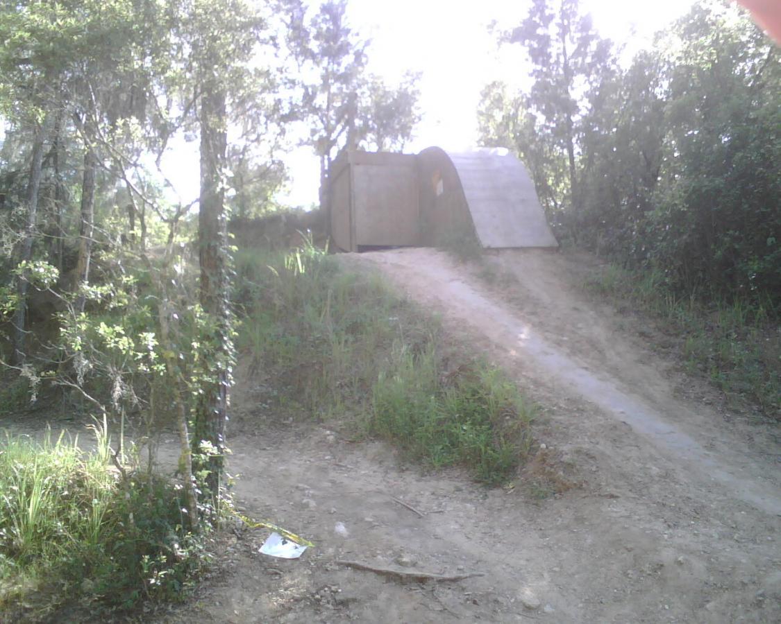 A dirt trail leading up to a curved wooden structure, surrounded by trees and greenery. The structure is situated on a slope, with sunlight filtering through the leaves above. Santos mountain bike trail.