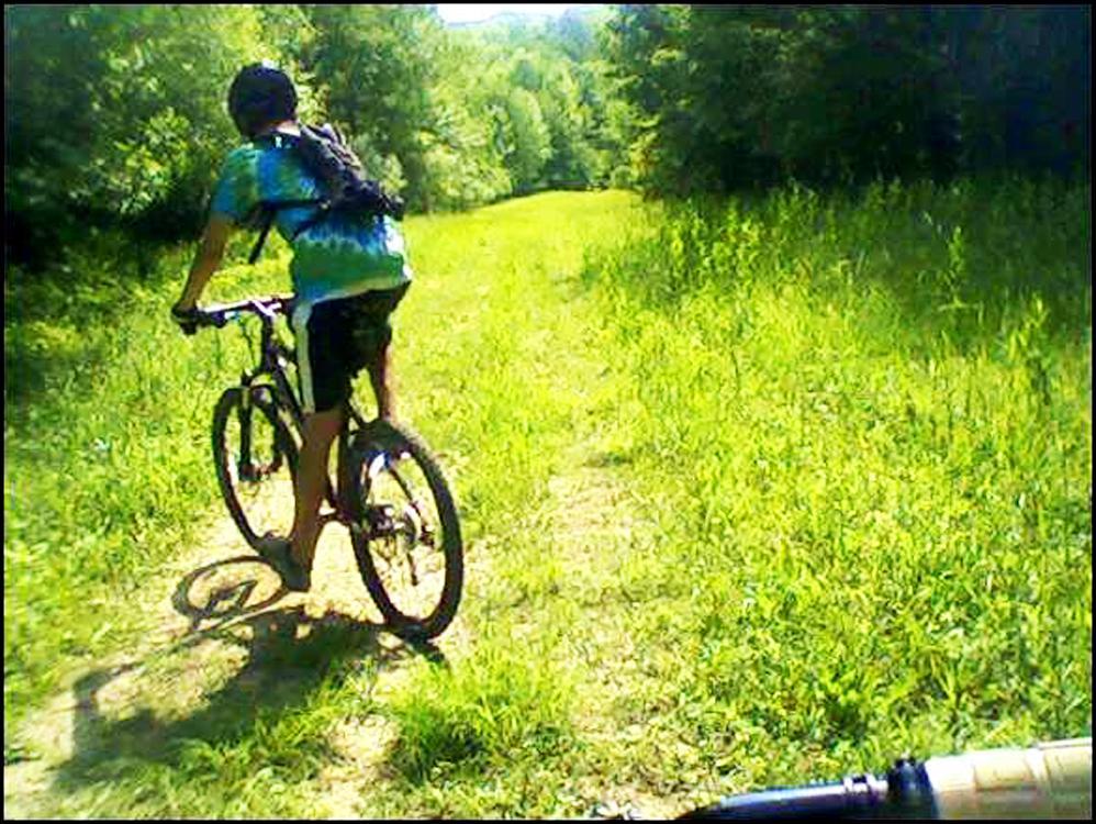 A person riding a mountain bike along a grassy path in a wooded area, with trees and greenery visible in the background. The individual is wearing a tie-dye shirt and shorts, and a backpack is secured to their back. Hunter's Creek Park Trails mountain bike trail.