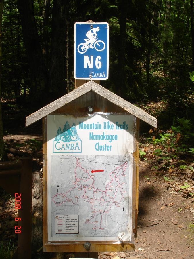 A wooden sign displaying a map of mountain bike trails in the Namakagon Cluster, with a blue sign marked "N6" above it. The map indicates various trails and includes a legend for navigation. The surrounding area features dense greenery typical of a forest setting. Rock Lake mountain bike trail.