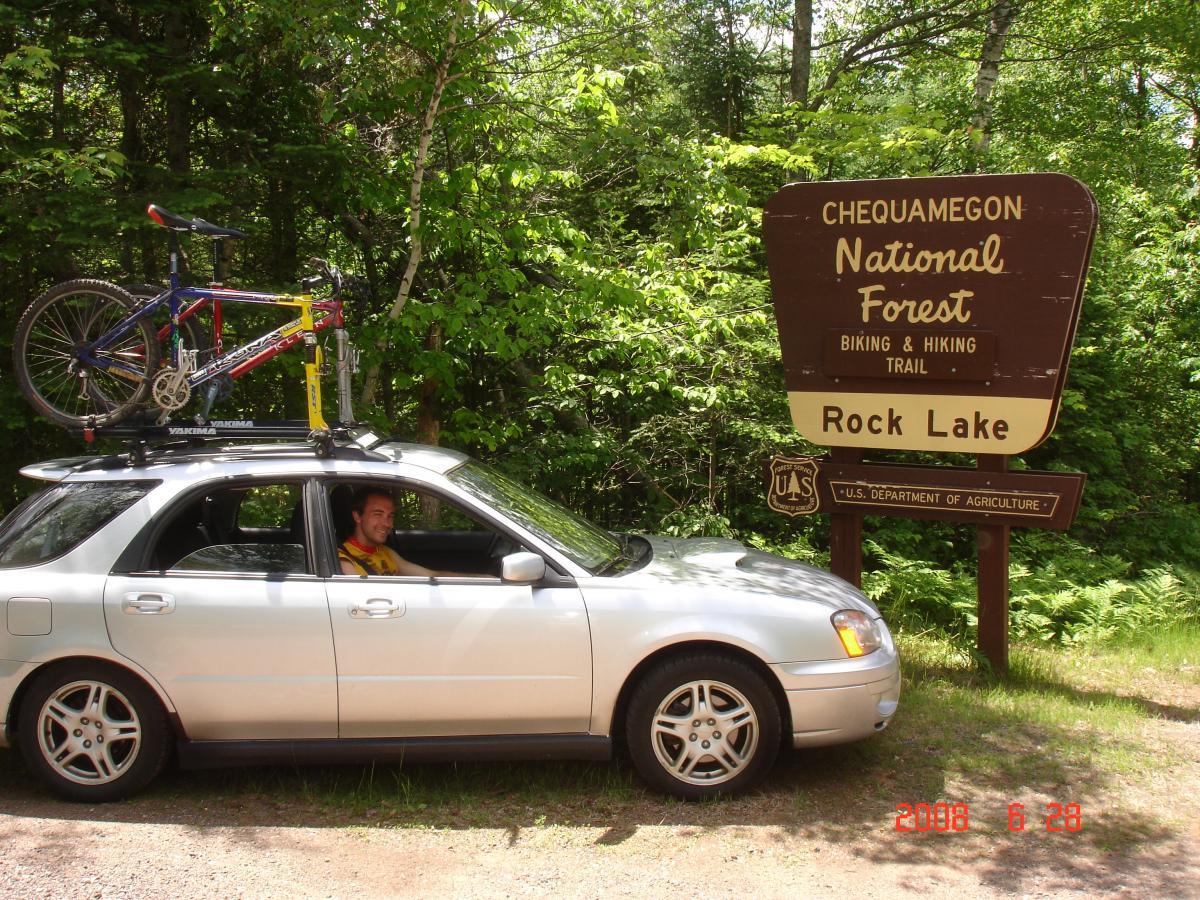 A silver car parked near a sign for Chequamegon National Forest, indicating a biking and hiking trail to Rock Lake. A colorful bicycle is secured on the roof of the car. A man sits in the driver's seat, smiling, with lush green trees in the background. The image is taken on a sunny day. Rock Lake mountain bike trail.