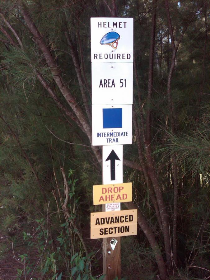 Signage posted along a wooded path, featuring four distinct signs: 

1. A sign indicating "Helmet Required" with an illustration of a helmet.
2. A sign labeled "AREA 51."
3. An "Intermediate Trail" sign with a blue square symbol and an upward arrow.
4. A sign stating "DROP AHEAD," followed by another sign for the "Advanced Section." 

The signs are surrounded by dense greenery. Oleta River State Park mountain bike trail.