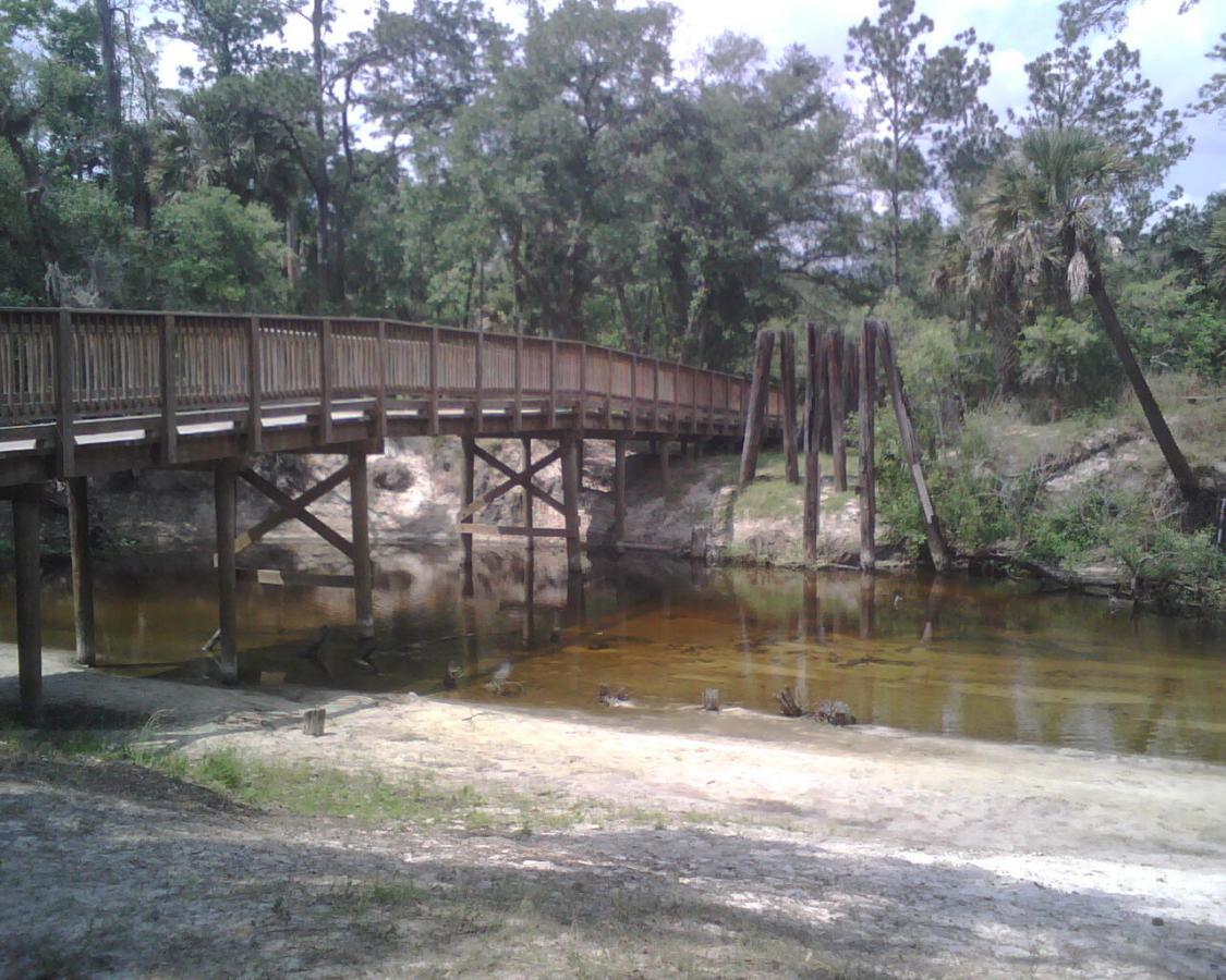A wooden bridge arches over a calm, shallow river, surrounded by lush greenery and tall trees. The river reflects the natural landscape, while sandy shores are visible in the foreground, creating a peaceful outdoor scene. Little Big Econ State Forest mountain bike trail.