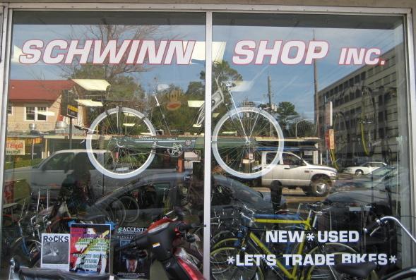 A storefront display for Schwinn Shop Inc. featuring a prominent bicycle in the window. The shop advertises both new and used bikes, with a message encouraging customers to trade bikes. Various bicycles are visible through the glass, alongside colorful promotional materials.