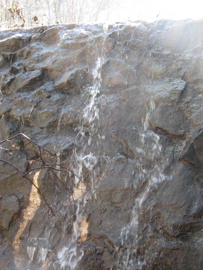 A close-up view of water cascading down a rocky surface, with sunlight reflecting off the wet stones. The surrounding area appears natural and untouched, with some twigs and branches visible among the rocks. Harris Shoals mountain bike trail.