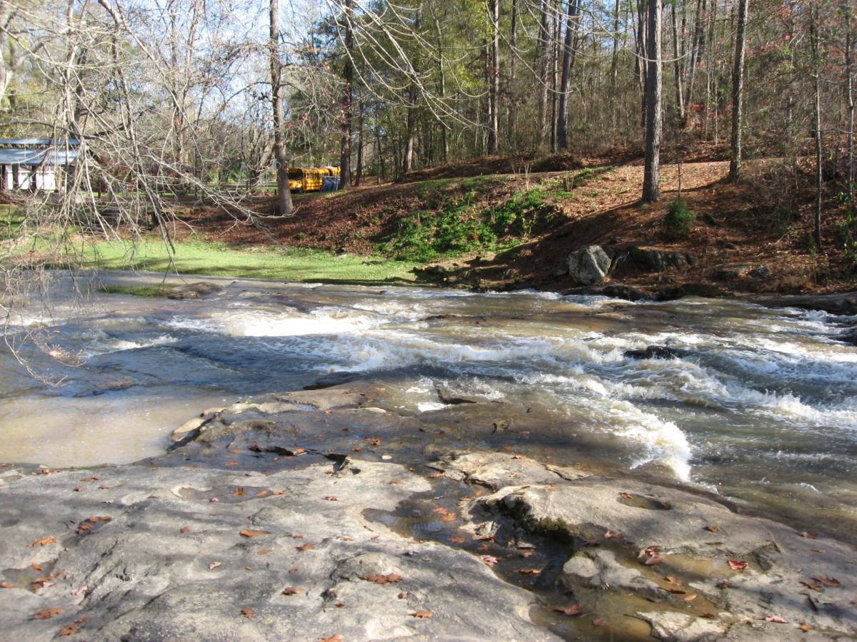 A peaceful river flows over rocky terrain, surrounded by trees and greenery. In the background, a glimpse of a building and a yellow vehicle can be seen, suggesting a nearby structure. The scene is framed by a clear blue sky, creating a serene natural environment. Harris Shoals mountain bike trail.