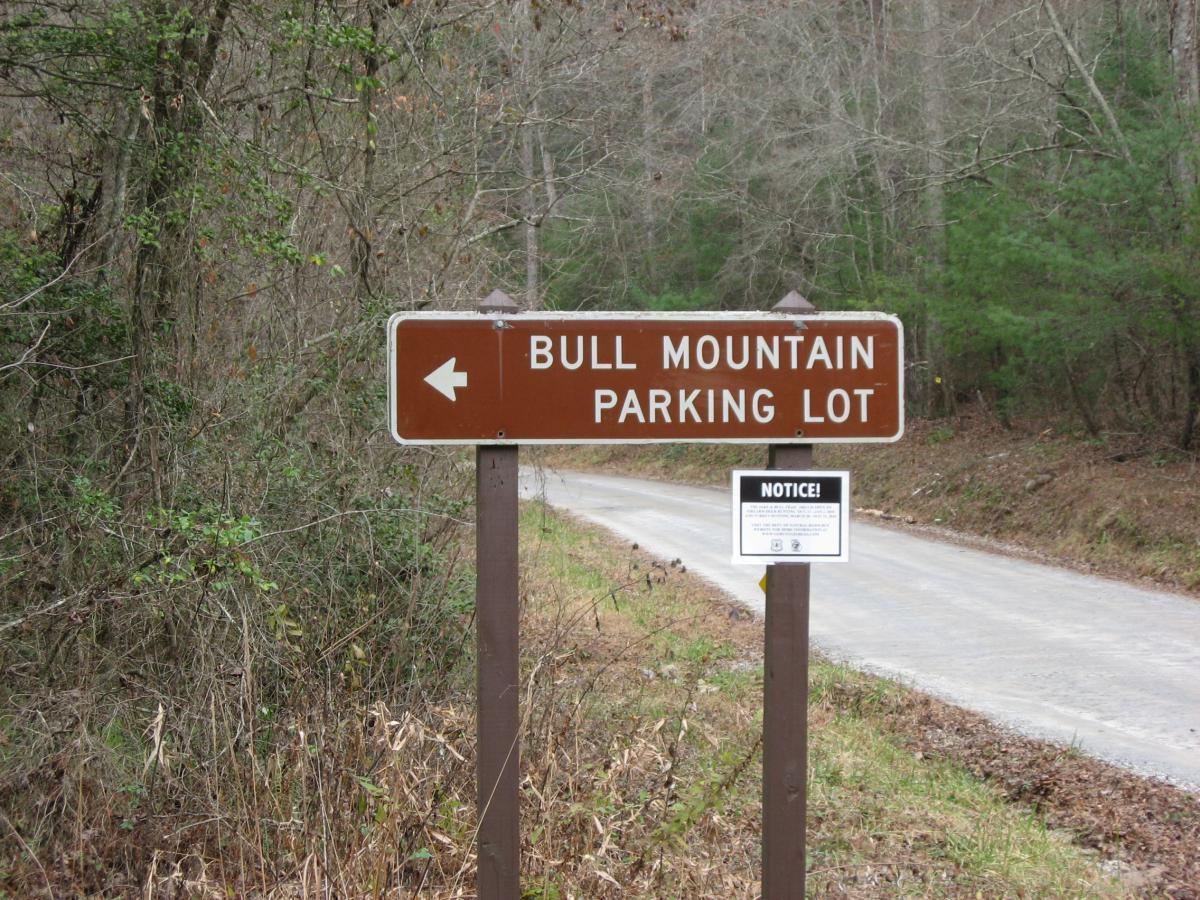 A brown sign indicating the direction to "Bull Mountain Parking Lot," with an arrow pointing left. Below the main sign, there is a smaller notice sign. The background features a tree-lined dirt road surrounded by greenery. Jake to Bull Mountain Connecter mountain bike trail.