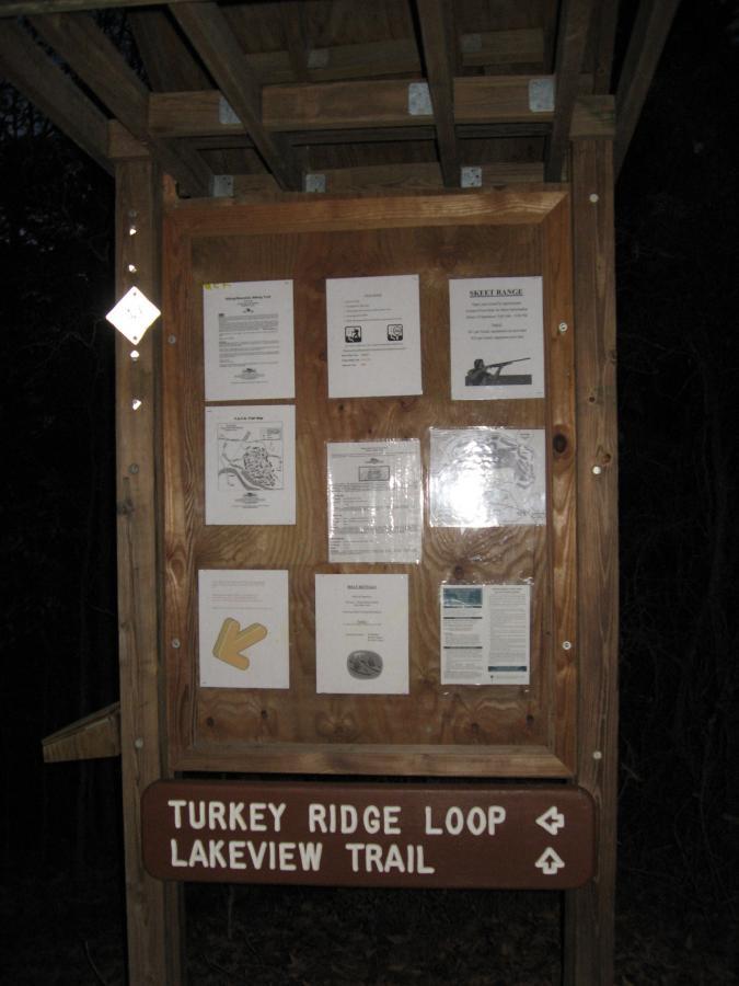 Wooden trail signboard displaying various informational flyers and maps, with directions to "Turkey Ridge Loop" and "Lakeview Trail" visible at the bottom. The setting appears dim, suggesting it may be evening or twilight. Hickory Knob State Park mountain bike trail.