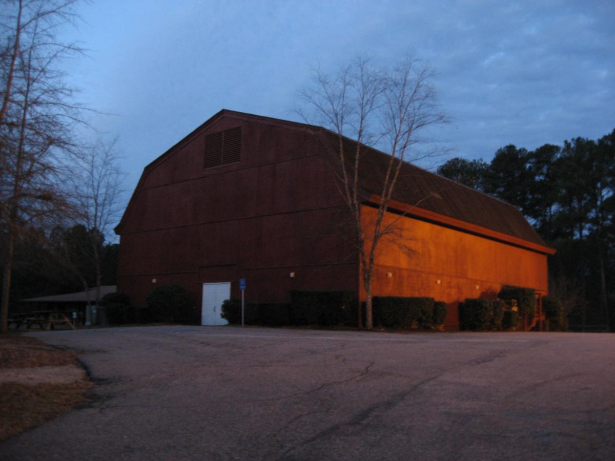 A large, barn-style building set against a dusk sky, with soft shadows and warm lighting on its exterior. The foreground features an empty parking area and a few bare trees, while a wooded area is visible in the background. A white door is prominent in the building's wall. Hickory Knob State Park mountain bike trail.