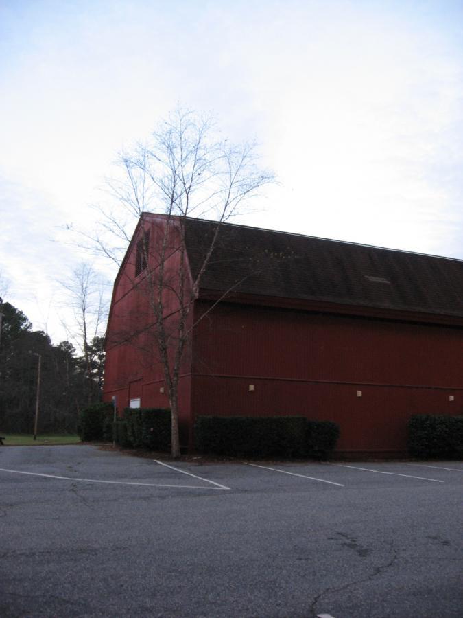 A large red barn stands in a parking lot, with a few sparse trees nearby and a cloudy sky in the background. The area is mostly empty, with visible parking spaces and a hedge along the building's side. Hickory Knob State Park mountain bike trail.