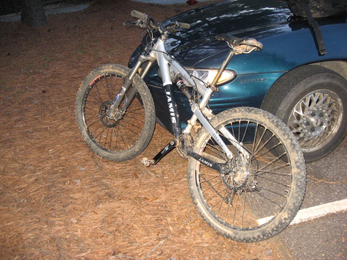 Alt text: A muddy mountain bike leaning against a parked car in a wooded area covered with pine needles. The bike has a silver frame, thick tires, and is positioned in a way that shows its front wheel and pedals. Hickory Knob State Park mountain bike trail.