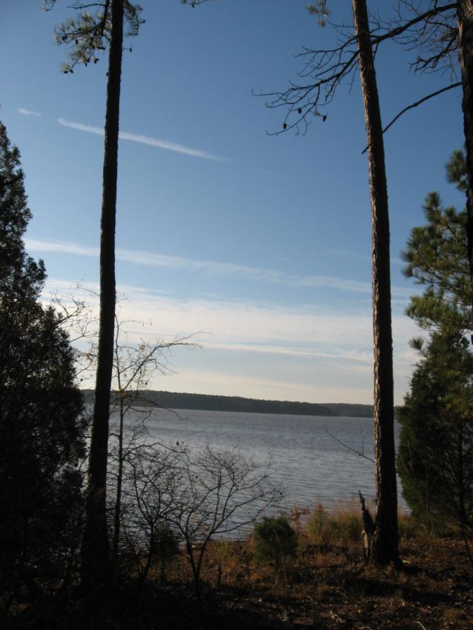 A serene view of a lake framed by tall trees against a clear blue sky, with gentle ripples on the water's surface and soft clouds in the background. Hickory Knob State Park mountain bike trail.