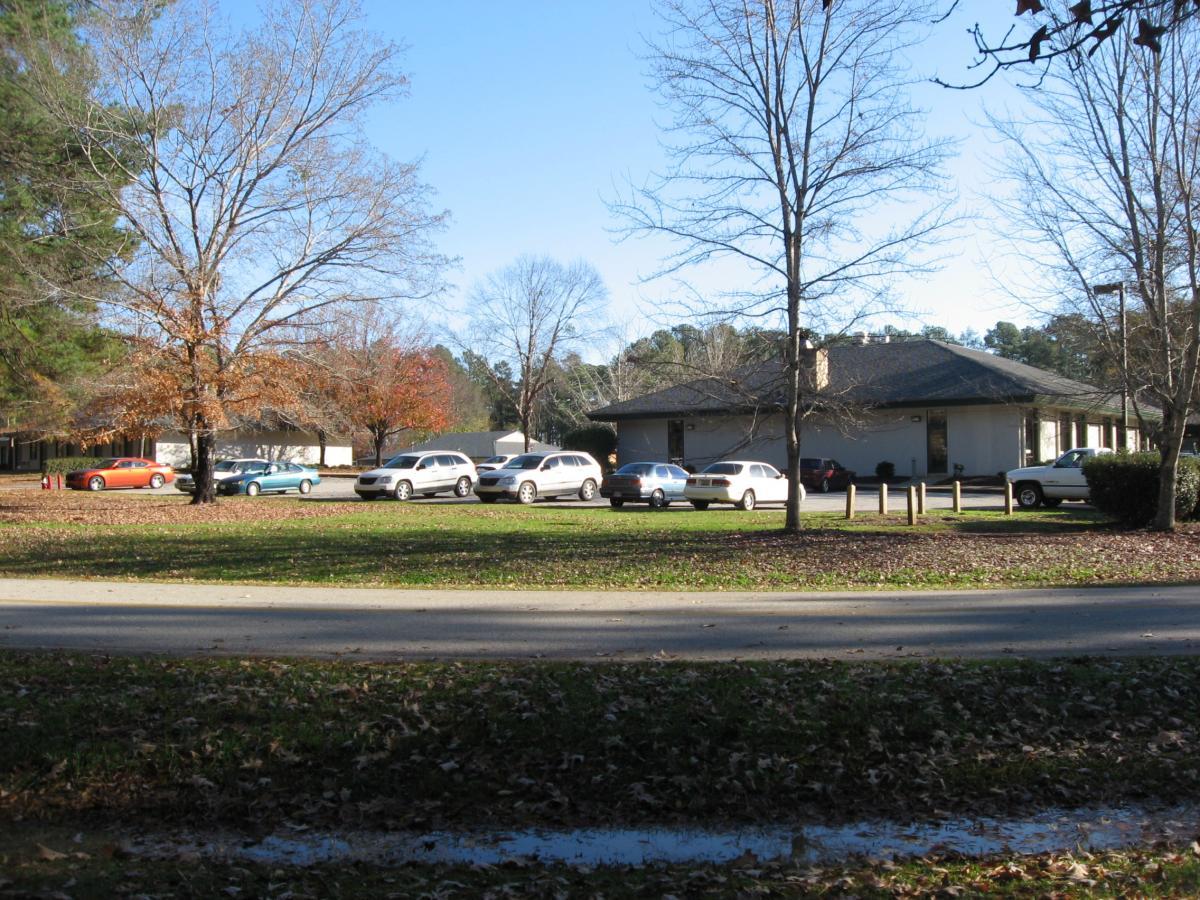 A view of a residential area featuring several parked cars, including a red vehicle and a teal car, along with a single-story building surrounded by trees. The scene is set under a clear blue sky, with a mix of deciduous trees showing bare branches and some remaining leaves. A street runs in the foreground, with fallen leaves scattered on the ground. Hickory Knob State Park mountain bike trail.