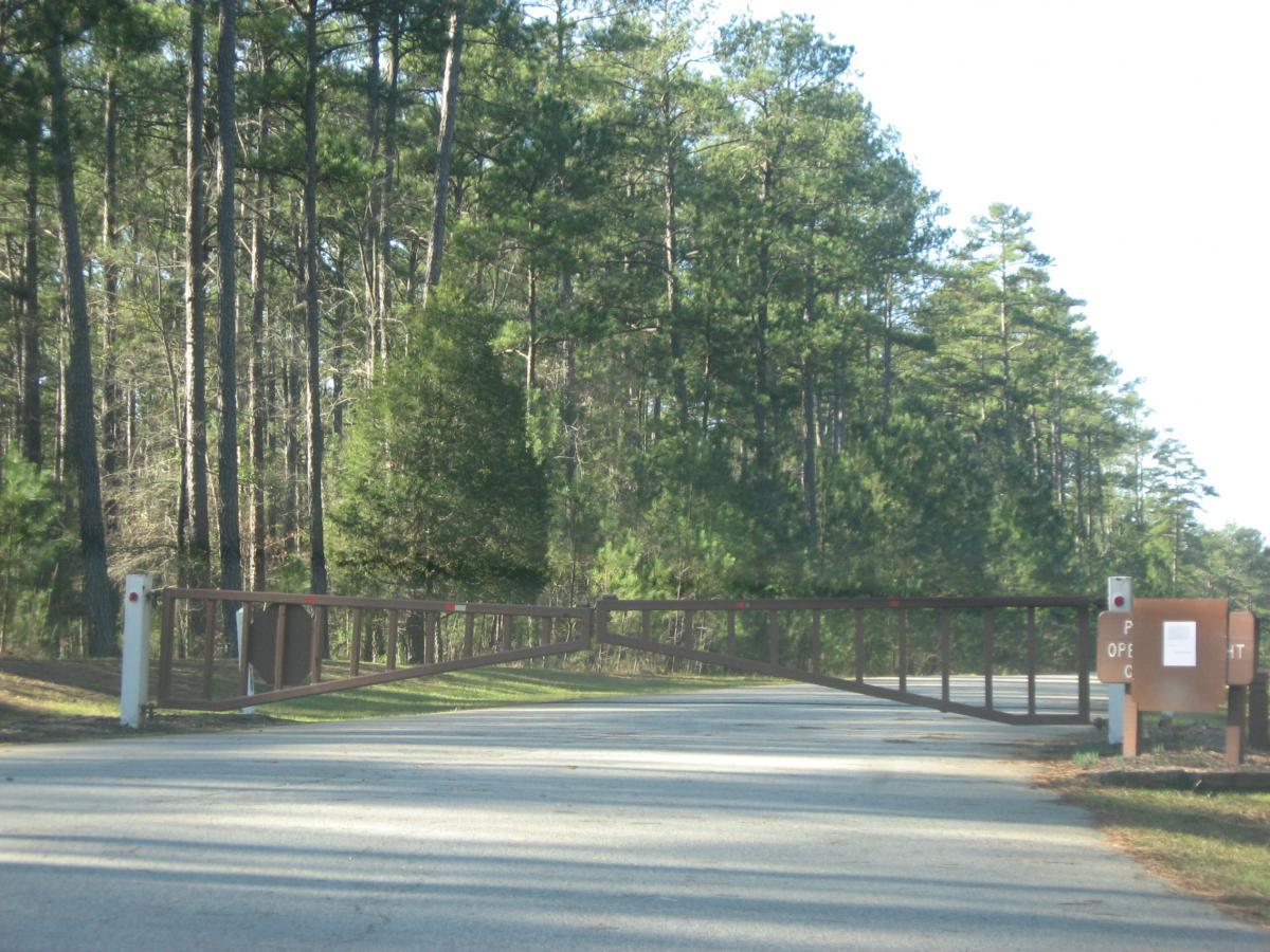A wooden gate spans a gravel road leading into a wooded area, surrounded by tall pine trees. Beside the gate, a sign indicates that the park is open for visitors. Sunlight filters through the trees, casting a serene atmosphere. Baker Creek mountain bike trail.