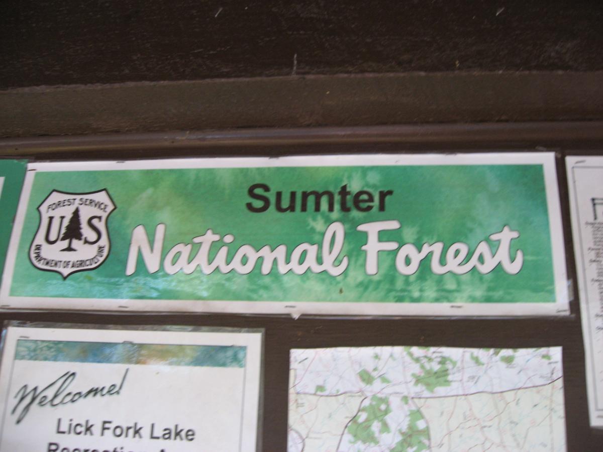 Sign for Sumter National Forest, featuring the U.S. Forest Service emblem. The background is green, and the text displays "Sumter National Forest" in large, bold letters. Additional informational materials are visible below the sign. Lick Fork (Horn Creek) mountain bike trail.