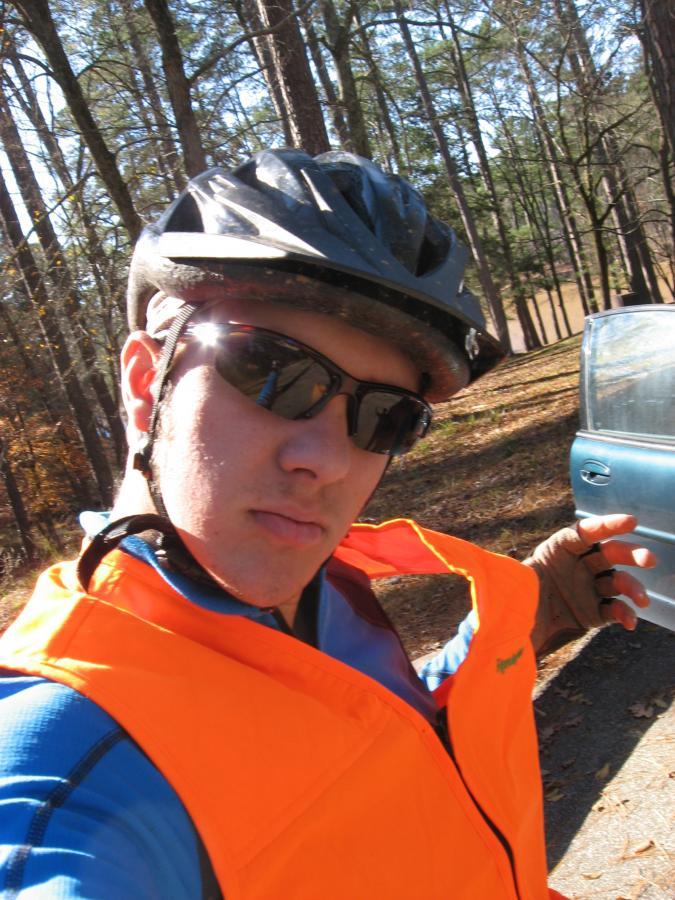 A young person wearing a black bicycle helmet and sunglasses is taking a selfie. They are dressed in a bright orange safety vest and a blue shirt. In the background, there are tall trees and a glimpse of a vehicle. The scene is outdoors, likely in a wooded area. Lick Fork (Horn Creek) mountain bike trail.