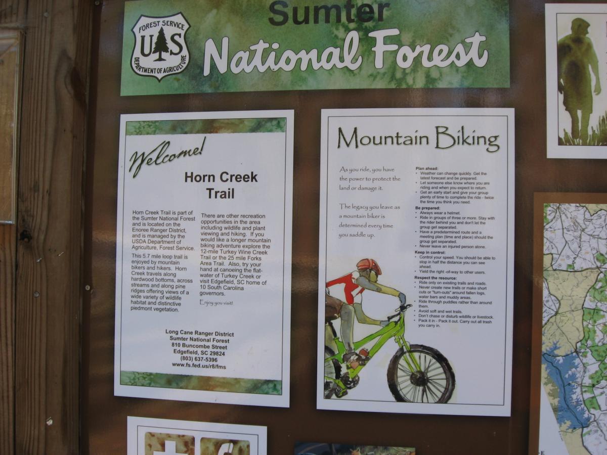 Alt text: Informational sign at Sumter National Forest featuring details about the Horn Creek Trail and guidelines for mountain biking. The sign includes a welcome message, trail description, safety tips, and a map. An illustration of a mountain biker is also present. Lick Fork (Horn Creek) mountain bike trail.