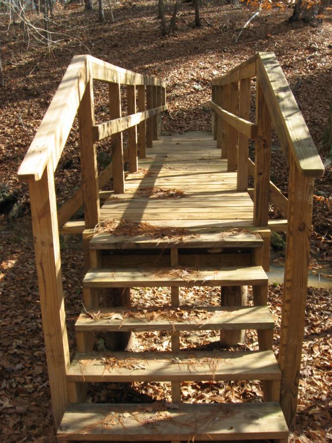 A wooden footbridge with a handrail spans a small creek, surrounded by a wooded area. The steps are adorned with fallen leaves, and sunlight filters through the trees, creating a warm atmosphere in the scene. Lick Fork (Horn Creek) mountain bike trail.