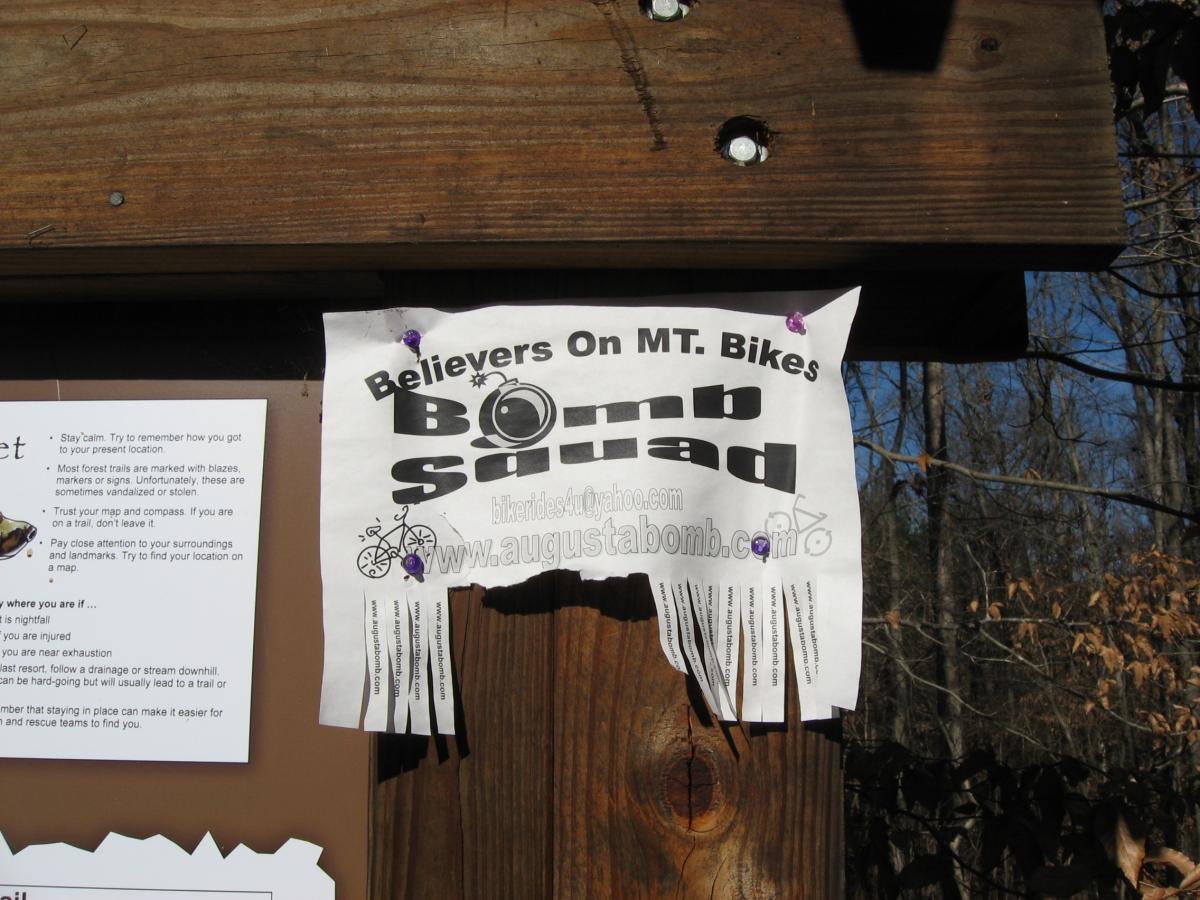 A poster titled "Believers On MT. Bikes" pinned to a wooden bulletin board in a forested area. The poster includes the name "Bomb Squad" and features tear-off strips at the bottom with website links. Nearby, there is another informational sheet outlining safety tips for outdoor activities, including directions for finding your location and what to do in various emergency situations. The setting is surrounded by trees, indicating a recreational area for mountain biking. Modoc (Stevens Creek Trail) mountain bike trail.