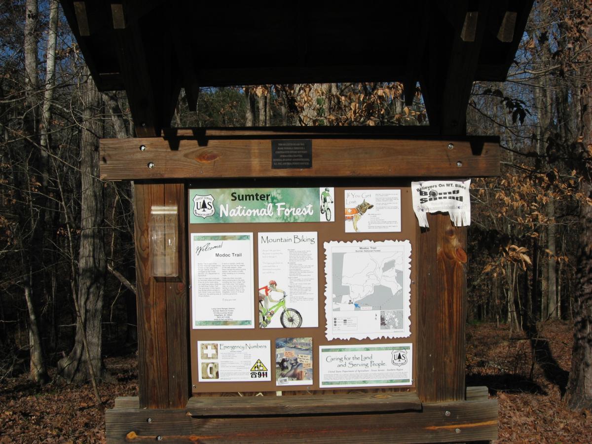 A wooden information board at Sumter National Forest displaying various notices and maps related to recreational activities, including mountain biking and the Modoc Trail. The backdrop features trees and natural surroundings typical of a forested area. Modoc (Stevens Creek Trail) mountain bike trail.