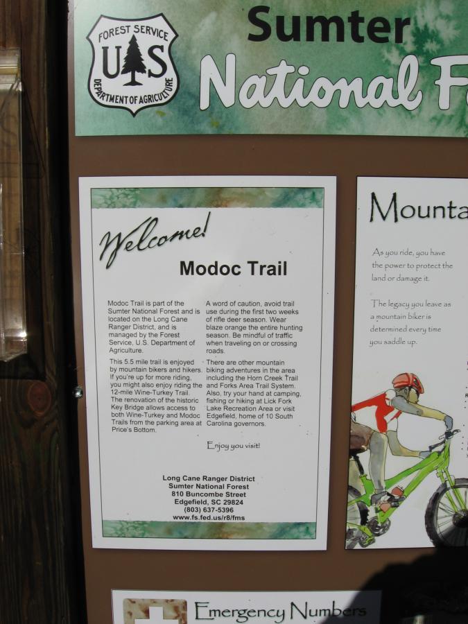 A bulletin board with information about the Modoc Trail in Sumter National Forest. The notice includes details on the trail's features, cautions for hikers, and contact information for the Long Cane Ranger District. Elements include a header with the U.S. Forest Service logo, a welcoming message, and emergency numbers on the side. Additionally, there is a graphic of a cyclist, emphasizing outdoor activities. Modoc (Stevens Creek Trail) mountain bike trail.