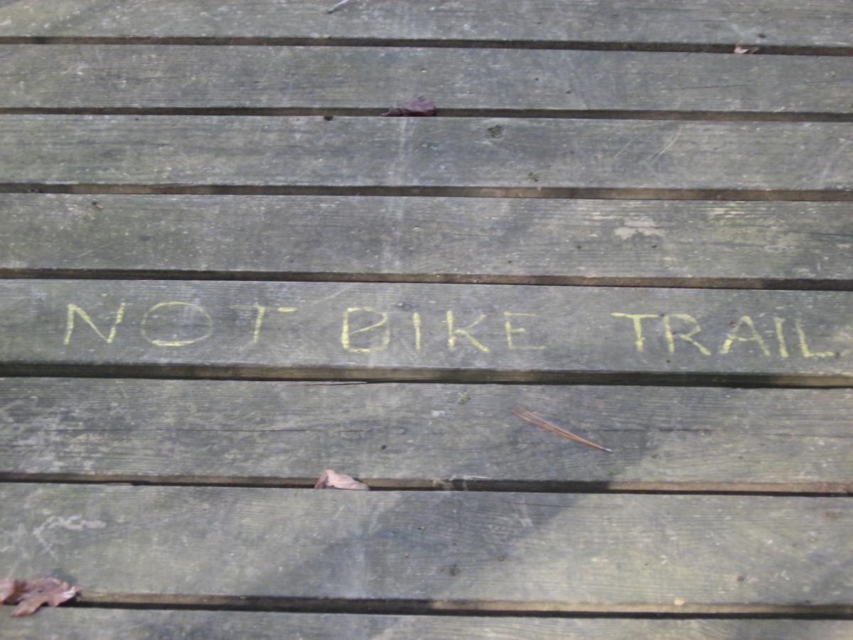 Wooden deck surface with the phrase "NOT BIKE TRAIL" written in yellow chalk. Some leaves are scattered on the planks. Modoc (Stevens Creek Trail) mountain bike trail.