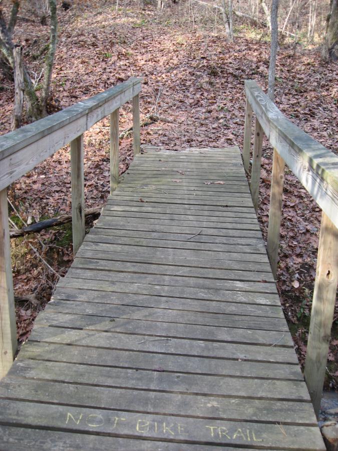 A wooden bridge leads over a small incline covered in fallen leaves in a wooded area. The bridge's edge is lined with wooden railings, and there is a message written in chalk on the bridge that reads "NOT BIKE TRAIL." Modoc (Stevens Creek Trail) mountain bike trail.