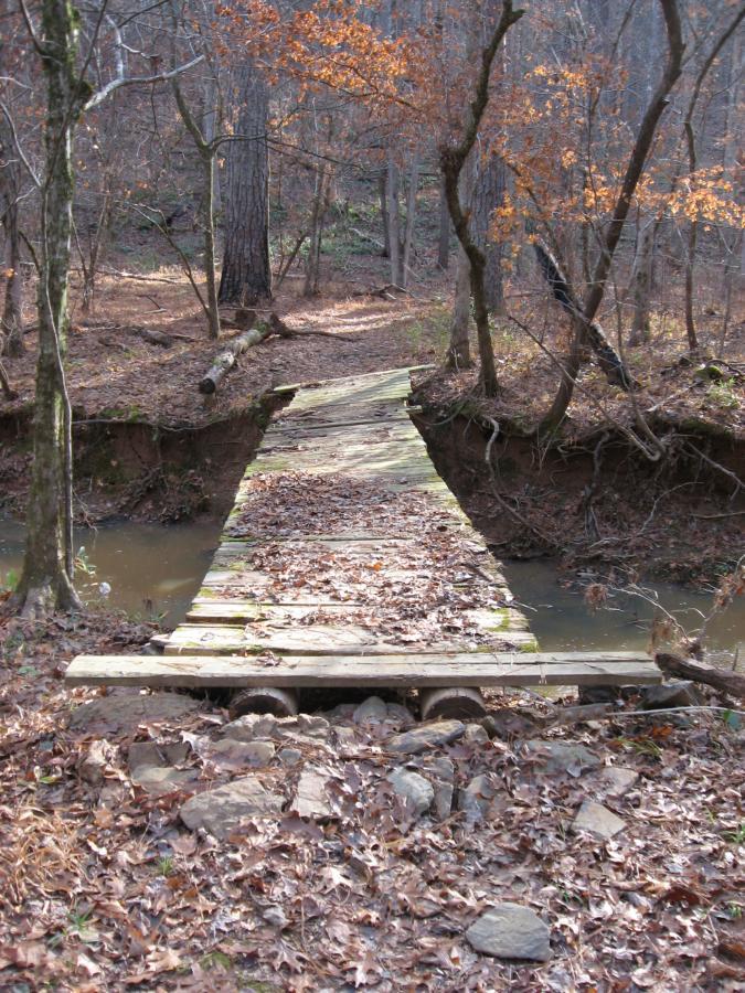 A rustic wooden bridge spans a narrow stream in a wooded area, surrounded by trees with sparse autumn leaves. The ground is covered in fallen leaves and rocks, indicating a quiet, natural setting. The bridge appears weathered and is partly supported by stones on either side. Modoc (Stevens Creek Trail) mountain bike trail.