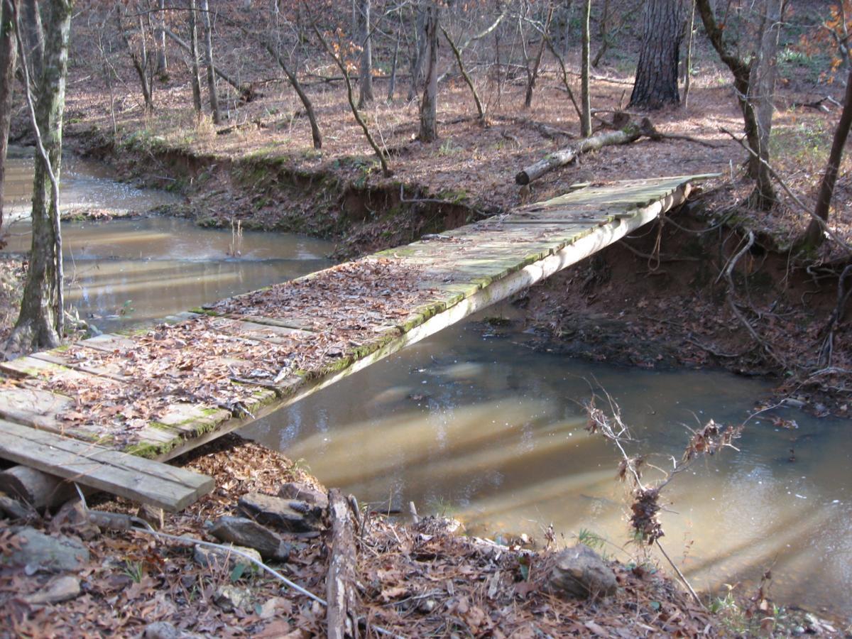 A rustic wooden footbridge crosses a shallow stream in a wooded area, surrounded by trees and fallen leaves. The water reflects the surrounding greenery and the sunlight filtering through the branches. Modoc (Stevens Creek Trail) mountain bike trail.