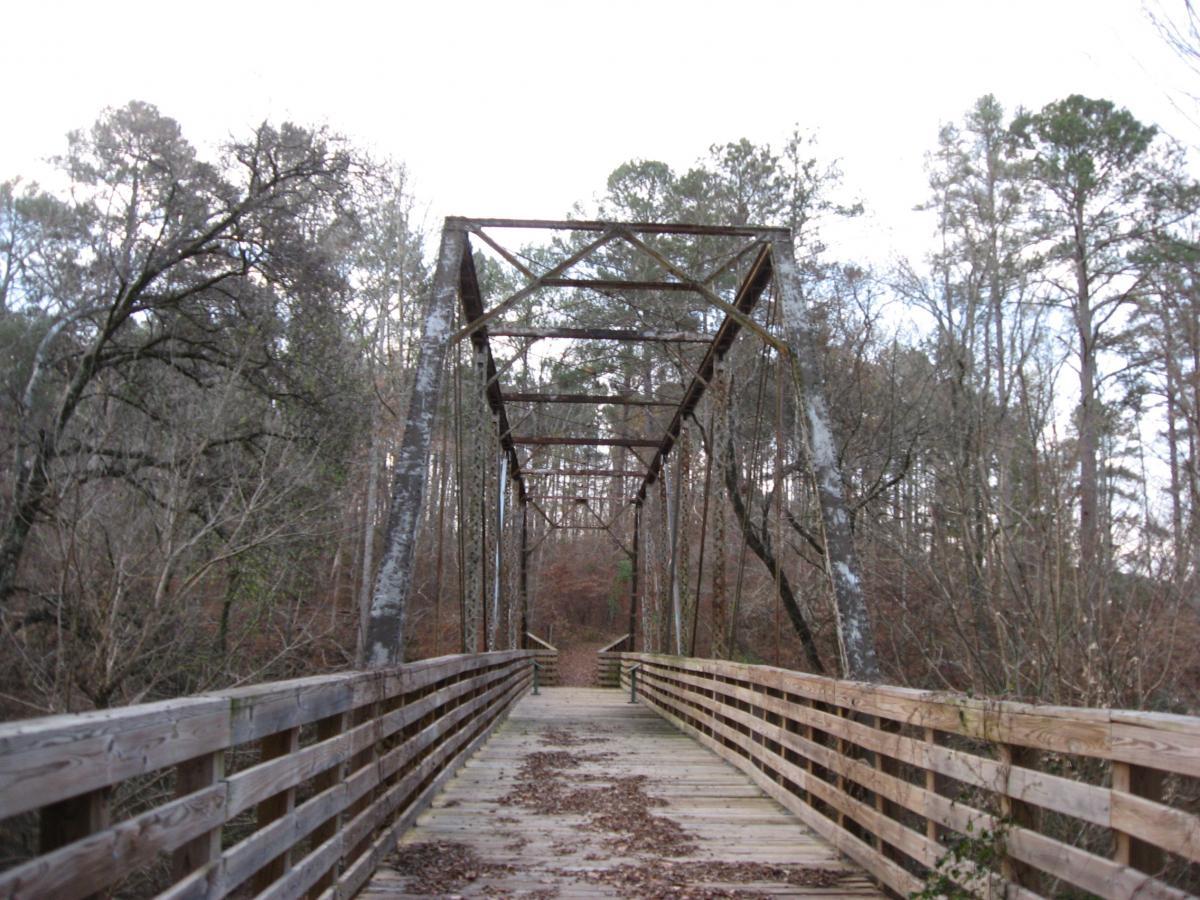 Old wooden bridge with a metal frame, surrounded by trees, leading over a path in a forested area. The bridge features a railing and is covered with leaves, indicating a natural, tranquil setting. Turkey Creek mountain bike trail.