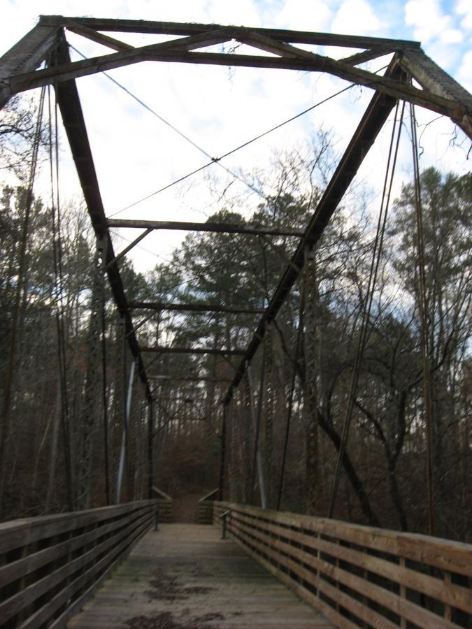 A wooden bridge with a metal frame and cables stretches into a wooded area, surrounded by trees. The sky is partly cloudy, and the bridge is empty, with wooden planks visible in the foreground. Turkey Creek mountain bike trail.