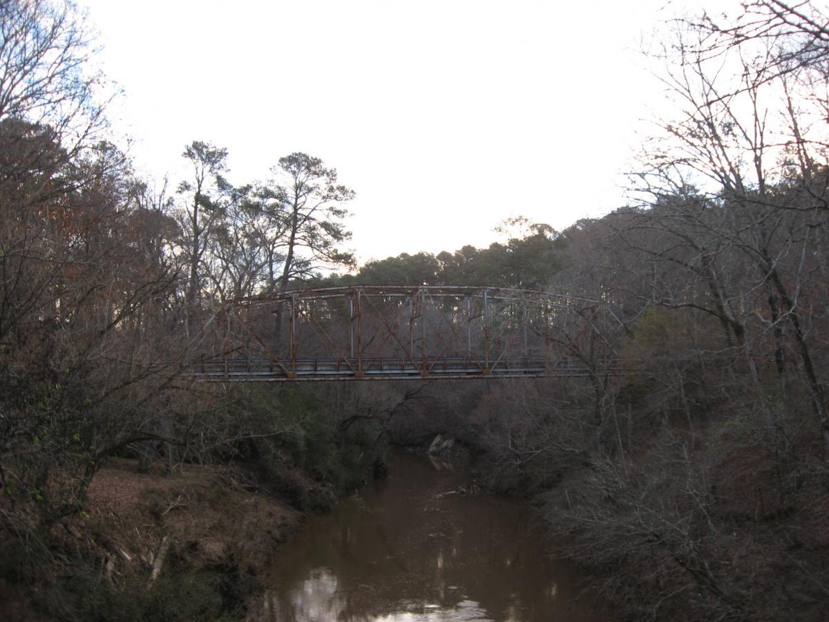 A weathered metal bridge spans a tranquil, winding creek, surrounded by bare trees and a landscape transitioning from autumn to winter. The sky above is soft and overcast, suggesting an early morning or late afternoon scene. Turkey Creek mountain bike trail.