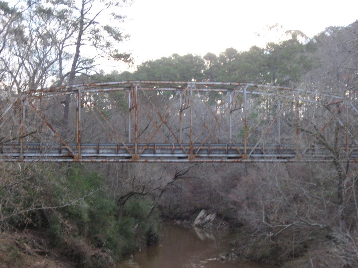 A rusty metal truss bridge spans a narrow, muddy river, surrounded by sparse trees and underbrush. The setting is quiet and rural, suggesting a sense of abandonment or disuse. The bridge structure is partially obscured by bare branches, highlighting the contrast between the industrial design and the natural landscape. Turkey Creek mountain bike trail.